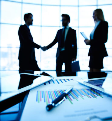 Business deal Technological devices, financial document with pen, glass of water at workplace on background of three business partners striking deal